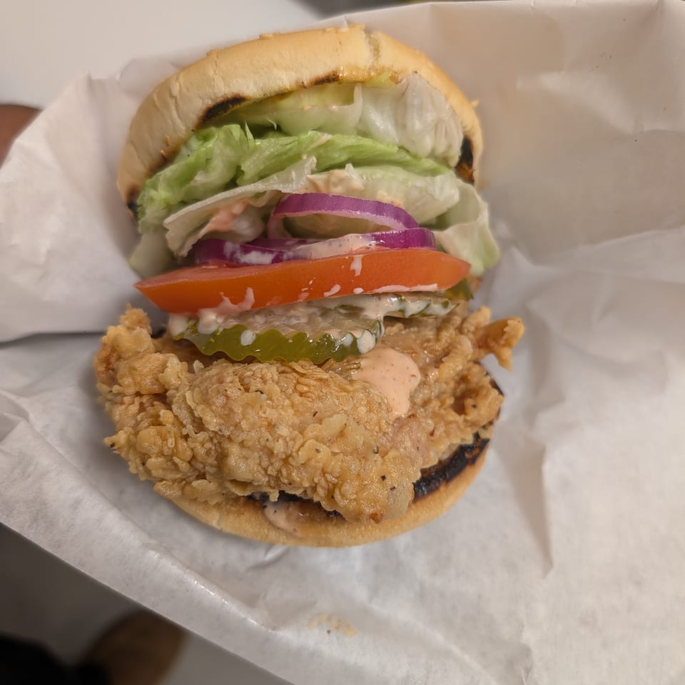 A Cap Hill fried chicken sandwich with lettuce, tomato, red onion, pickles, and sauce on a bun.