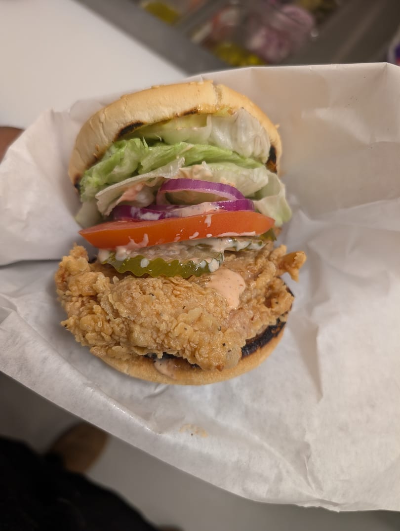 A Cap Hill fried chicken sandwich with lettuce, tomato, red onion, pickles, and sauce on a bun.