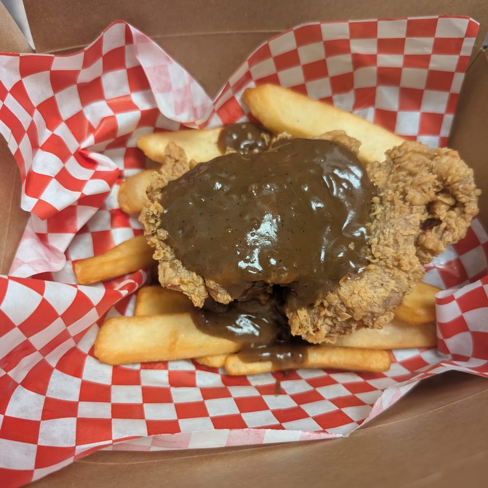 Fried chicken steak with gravy and fries in a takeout box with red and white checkered paper.