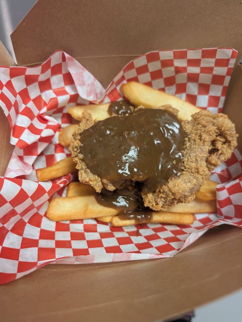 Fried chicken steak with gravy and fries in a takeout box with red and white checkered paper.